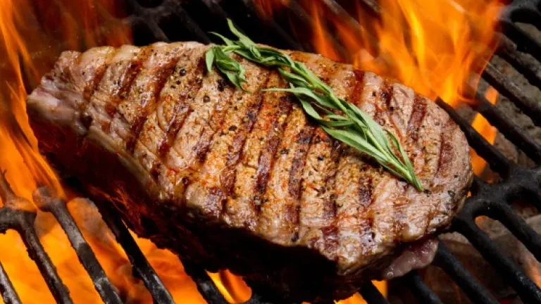 Thick grilled steak with rosemary, cooking over vibrant orange flames.