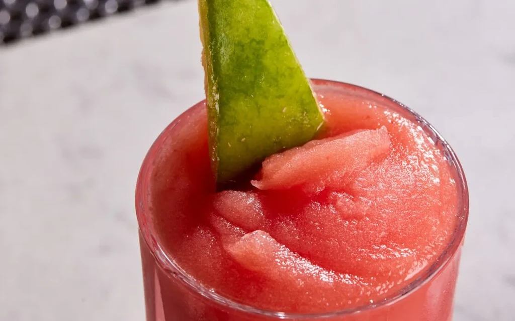 Vibrant red slushy drink in a glass, garnished with lime and fruit.