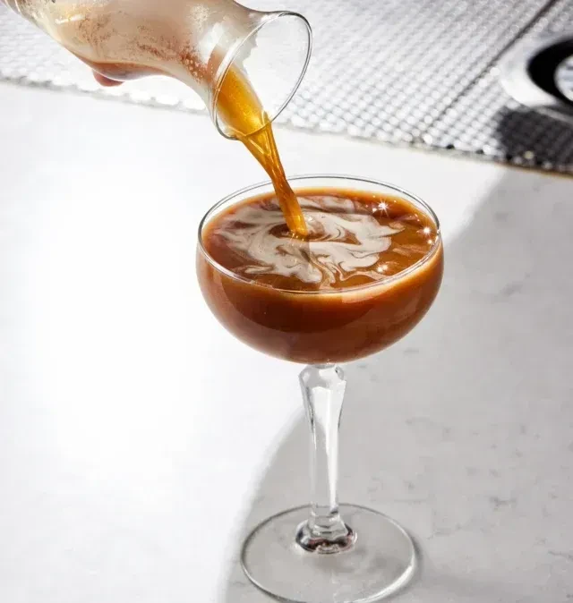 Hand pours golden liquid from pitcher into stemmed glass, creating foam.