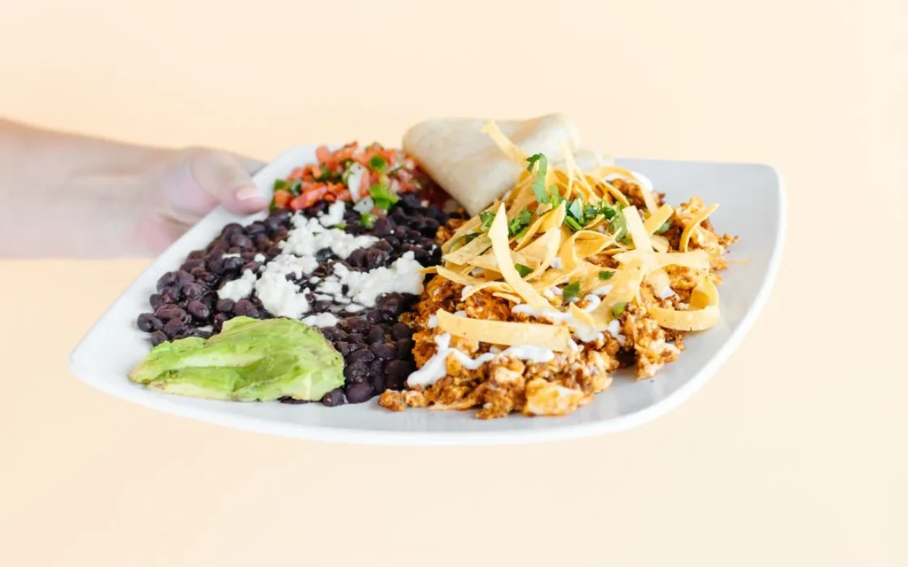 Hand holding a plate of Mexican-style breakfast with beans, eggs, and tortilla.