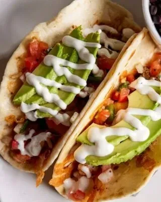 Two soft tacos topped with avocado, tomatoes, onions, and white sauce.