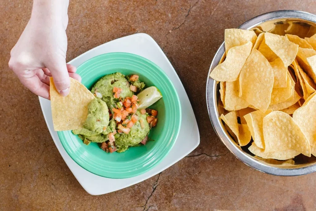 Hand dipping tortilla chip into guacamole, bowl of chips.