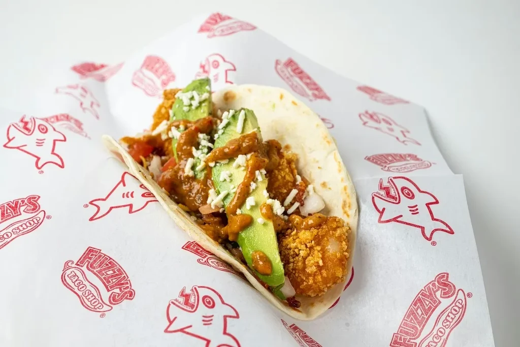 Fried seafood taco with avocado, cheese, and sauce on a branded wrapper.