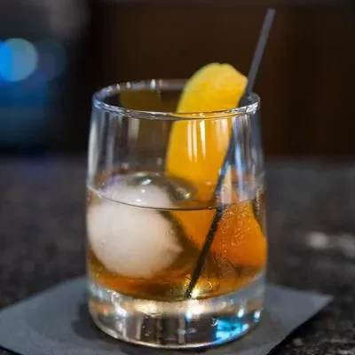 Amber cocktail in a tumbler with a spherical ice cube and citrus peel.