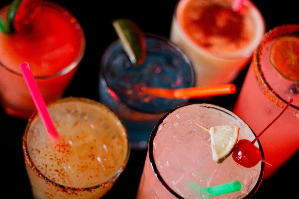 Six colorful garnished cocktails with rims, close-up on dark background.