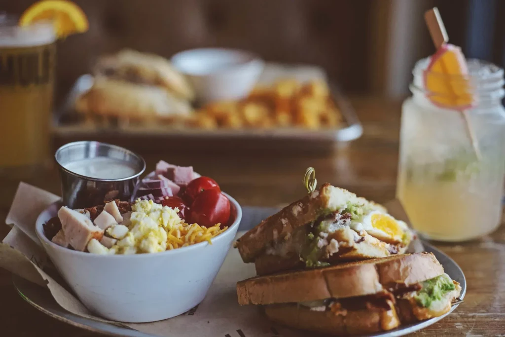 Colorful salad, club sandwich, and drinks on a rustic wooden table.