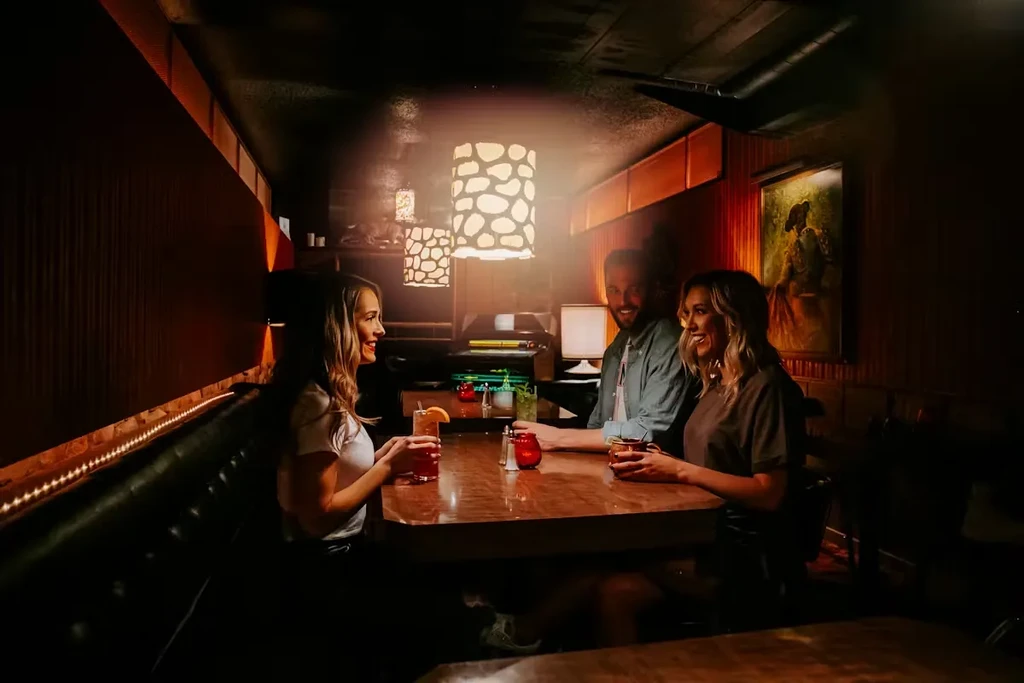 Two women and a man smiling, holding drinks in a cozy restaurant.