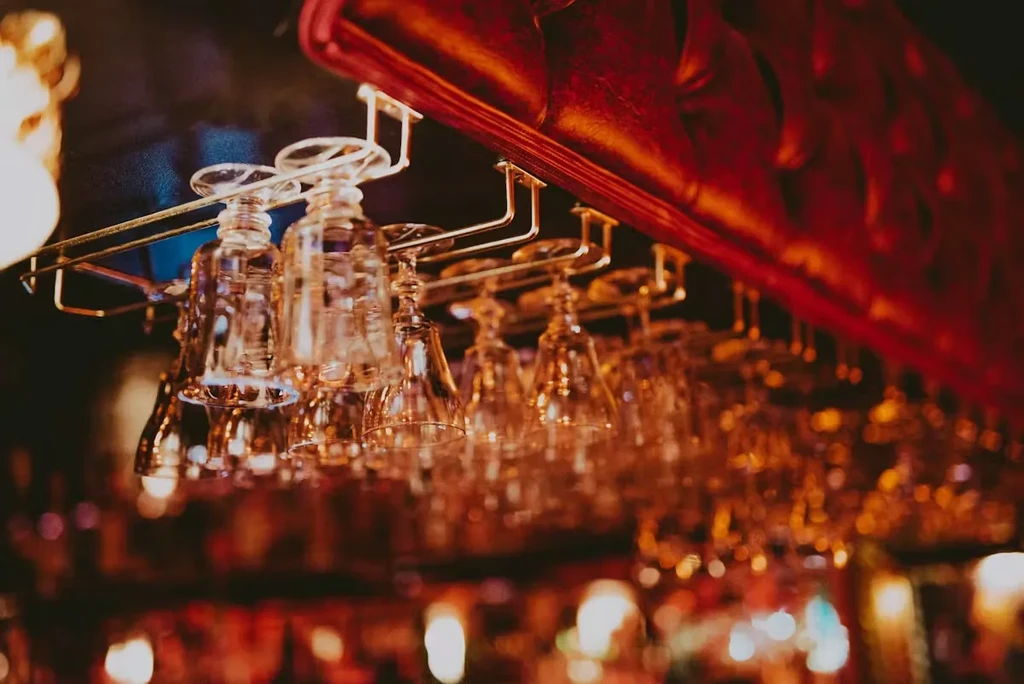 Clear wine glasses hang from a rack above a bar, with red upholstery.