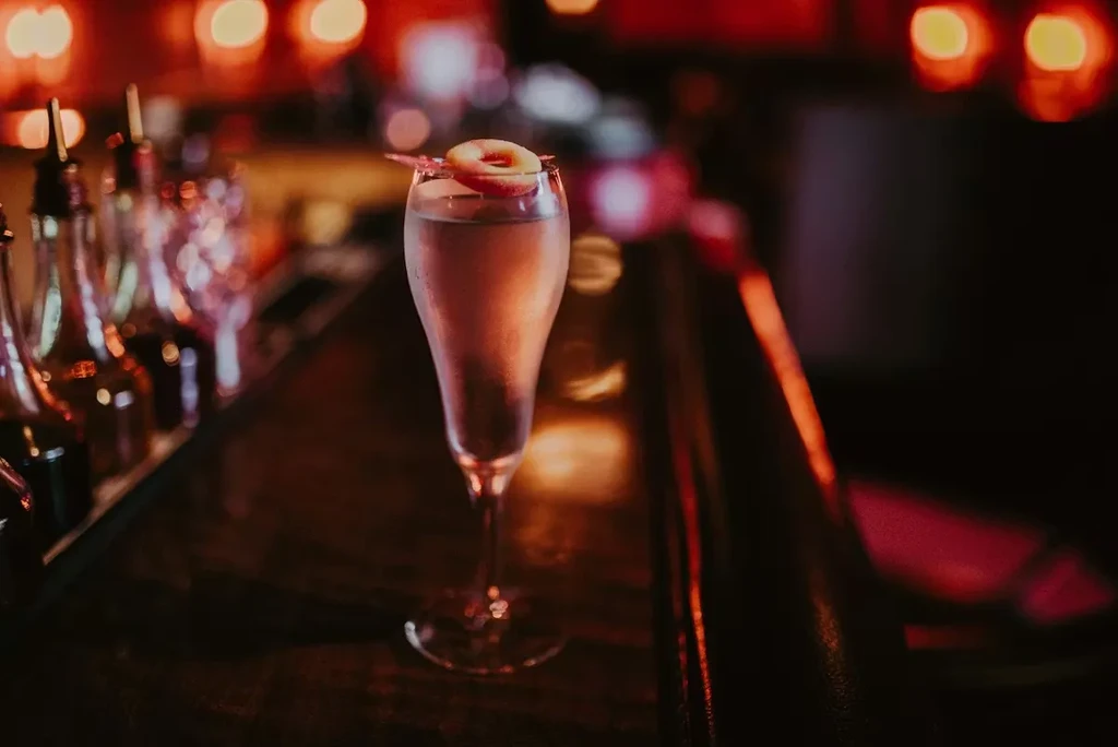 Cocktail in flute glass with fruit and pink garnish on bar.