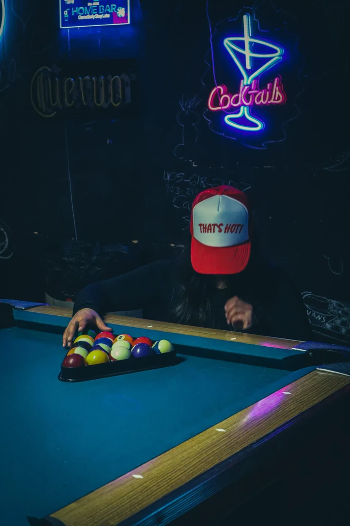 Person in 'THAT'S HOT!' hat racks pool balls in a dark bar.