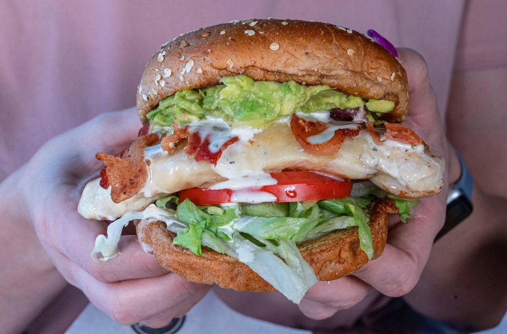 Tall grilled chicken sandwich with avocado and bacon, held by hands.