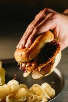 Hand holding large fried chicken sandwich, pickles, slaw, and chips.