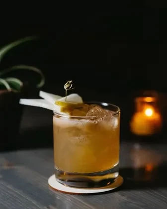 Close-up of an amber cocktail with ice and fruit garnish on a wooden surface.