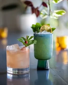 Two garnished cocktails, one orange/pink, one blue/green, on a dark surface.
