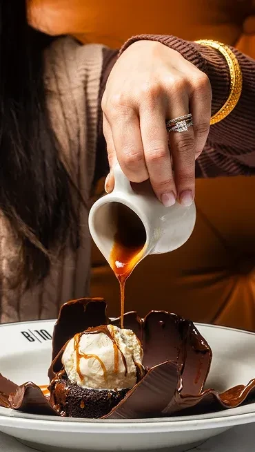 Hand pours amber sauce over chocolate dessert and vanilla ice cream.
