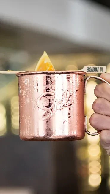 Hand holding a Stoli copper Moscow Mule mug with an orange garnish.