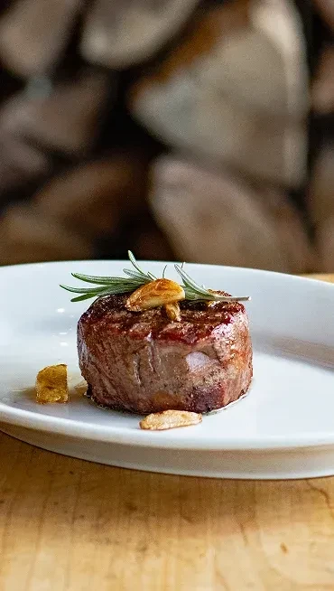 Cooked steak with rosemary and crispy garlic chips on a white plate.