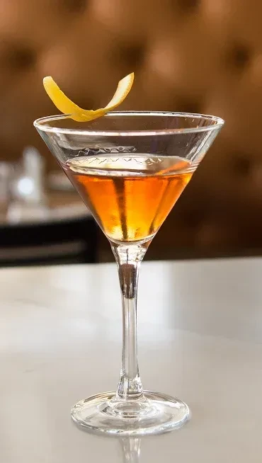 Amber cocktail in martini glass with curled yellow lemon peel.