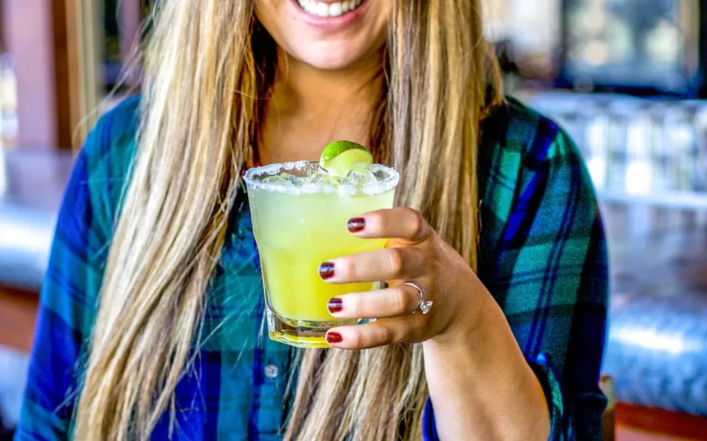 Smiling person holds a salt-rimmed green margarita with lime.