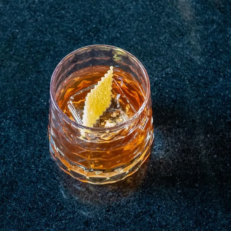 Amber cocktail in faceted rock glass with ice and lemon peel.