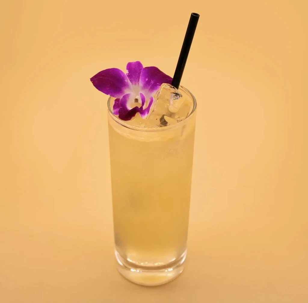 Golden cocktail in glass, ice, dark straw, purple orchid garnish.