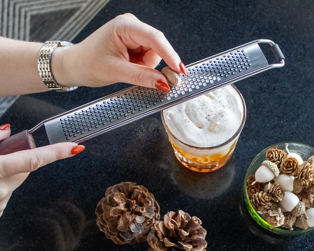 Person's hands grating nutmeg onto a foamy drink.