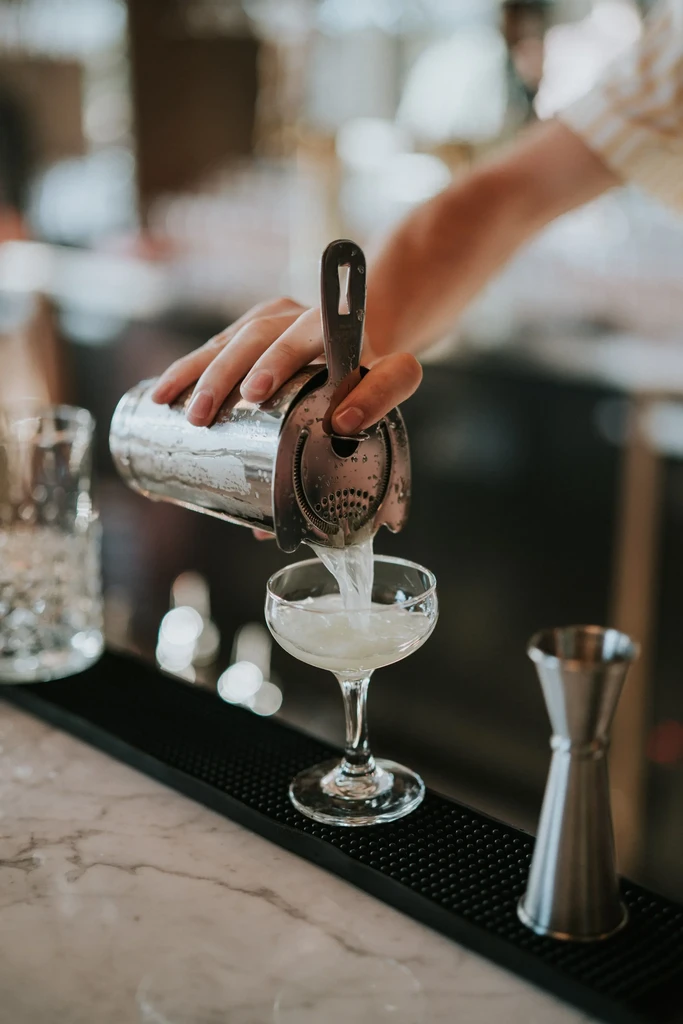 Bartender strains light cocktail from shaker into a coupe glass.