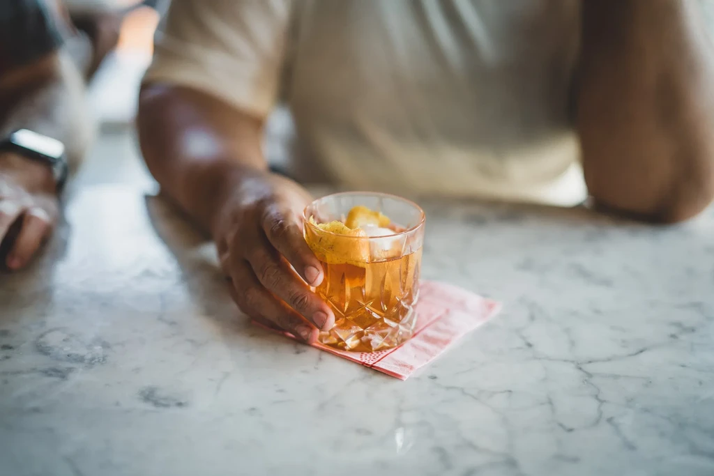 Hand holds amber cocktail with ice and citrus peel in cut glass.