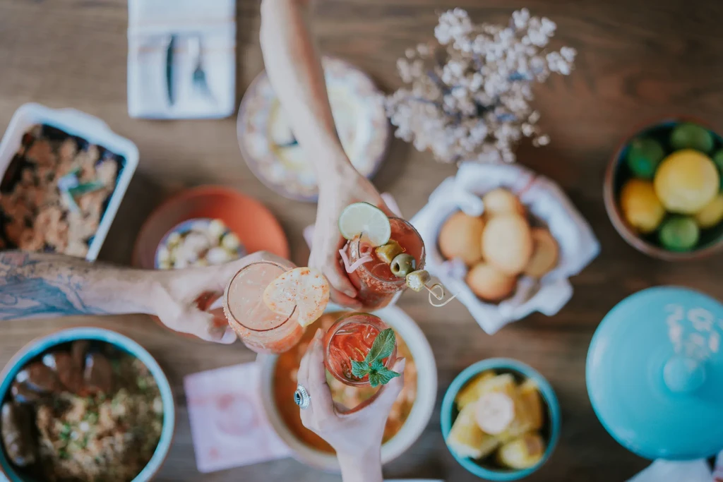 Hands clinking cocktail glasses over a dining table laden with food.