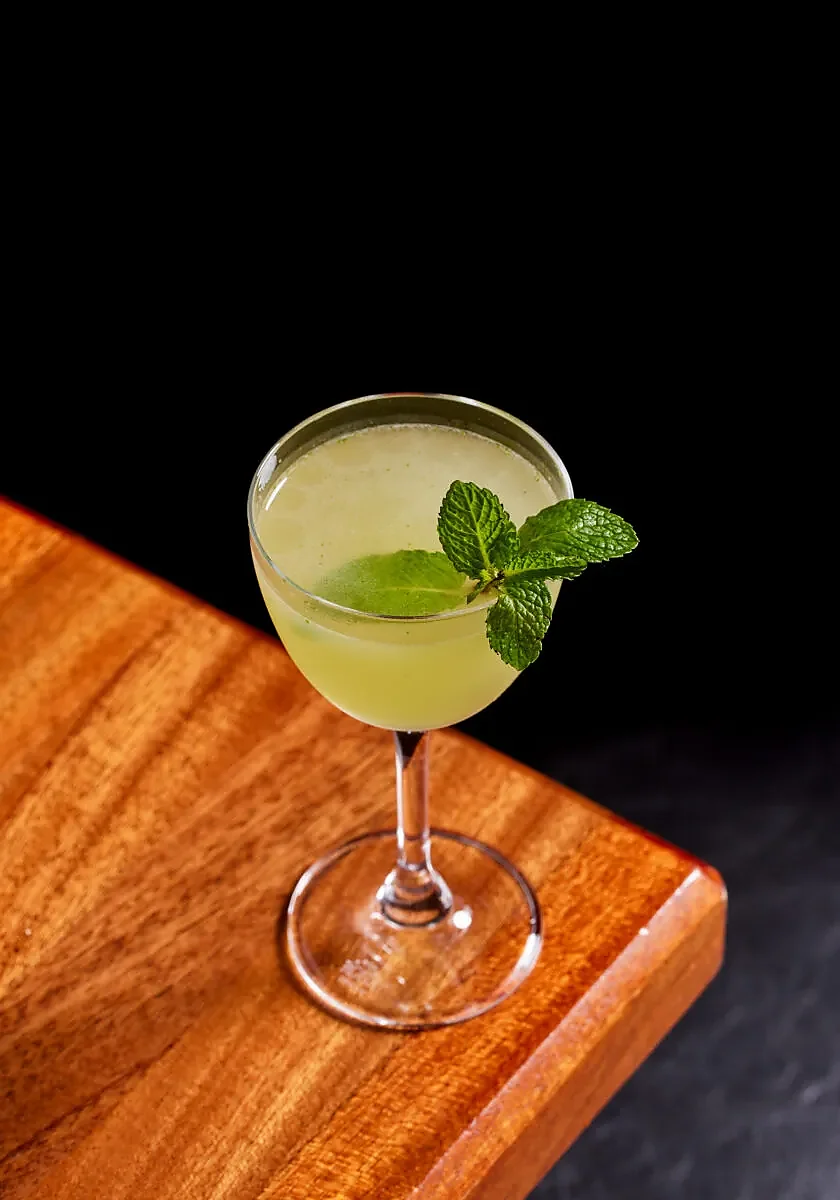 Pale green cocktail in stemmed glass with mint garnish on wood