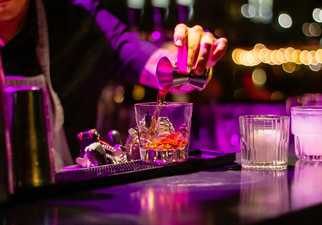 Bartender's hand pours golden spirit into icy glass, vibrant purple light.