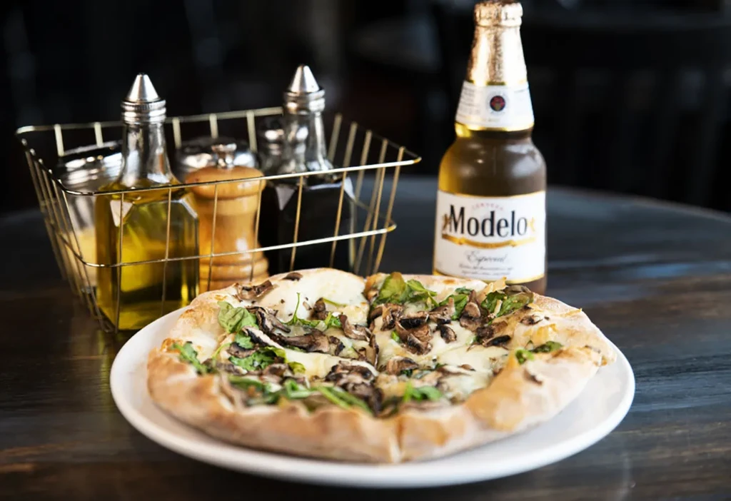 Gourmet pizza with mushrooms, arugula, and a Modelo beer on a dark table.