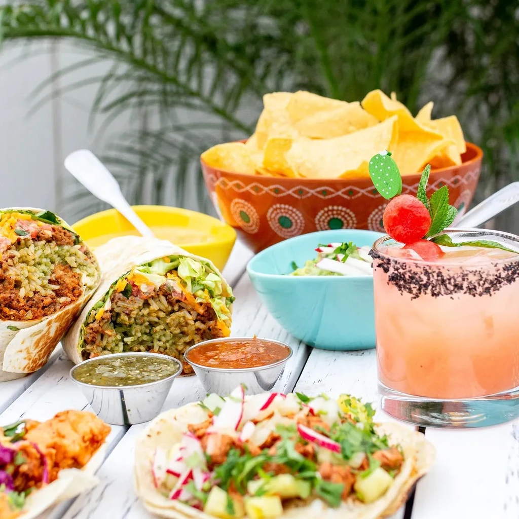 Vibrant Mexican food spread: burritos, tacos, chips, guacamole, cocktail.
