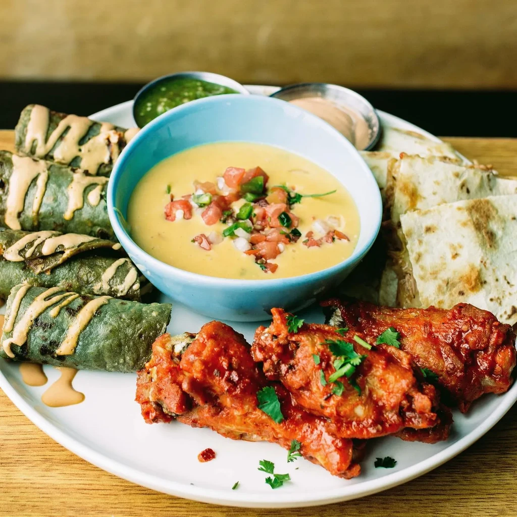 Food platter with queso dip, taquitos, sauced chicken wings, and quesadilla wedges.