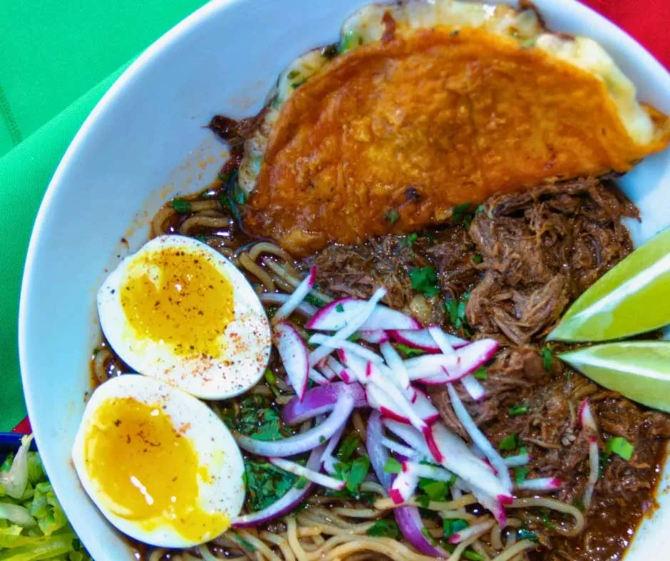 Overhead: Birria ramen bowl with quesabirria taco, soft-boiled egg, lime.