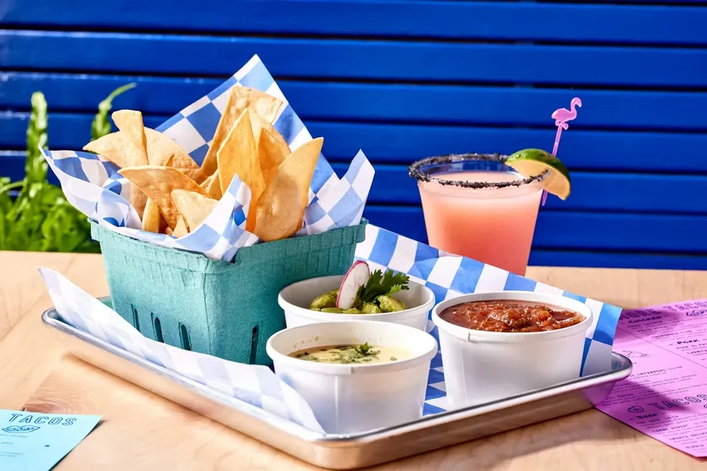 Tortilla chips, dips, pink cocktail on wood table with blue background.