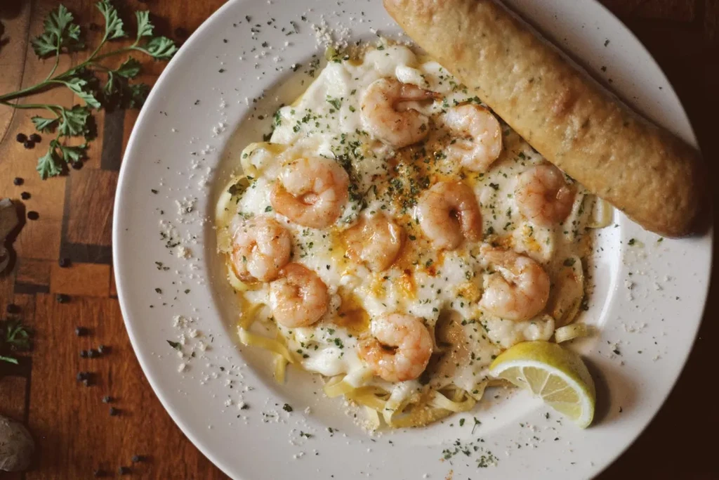 Overhead creamy shrimp pasta, breadstick, and lemon wedge on wood.
