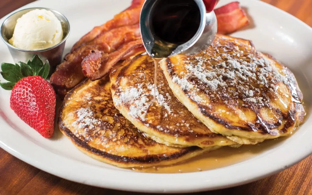 Pancakes with powdered sugar, pouring syrup, crispy bacon, and a strawberry.