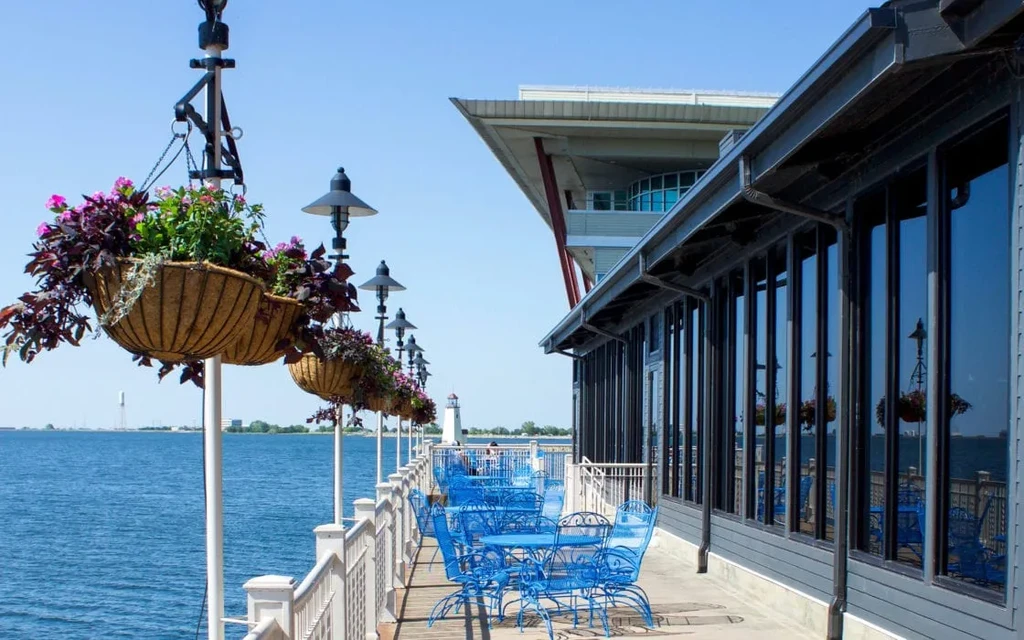 Sunny waterfront patio with blue tables and a distant lighthouse.