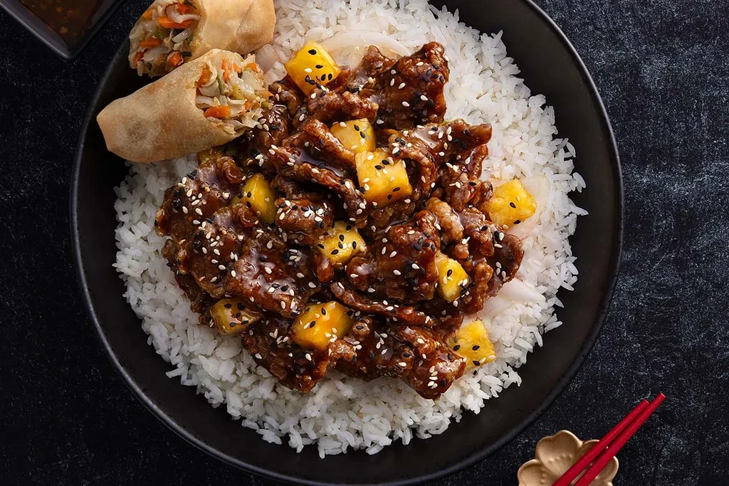 Top-down: Asian rice bowl with stir-fry, pineapple, spring rolls, chopsticks.