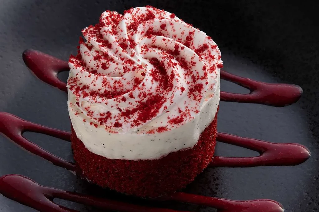 Red velvet dessert with cream cheese frosting and red sauce on dark plate.