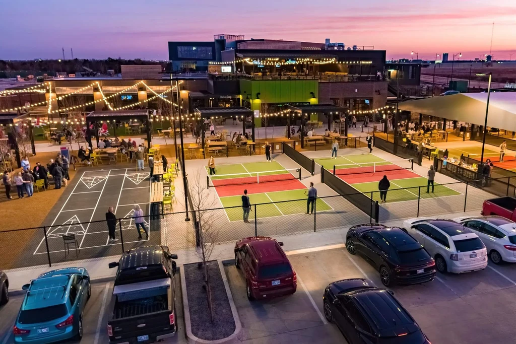 Aerial view of a lively outdoor complex with shuffleboard and pickleball at dusk.
