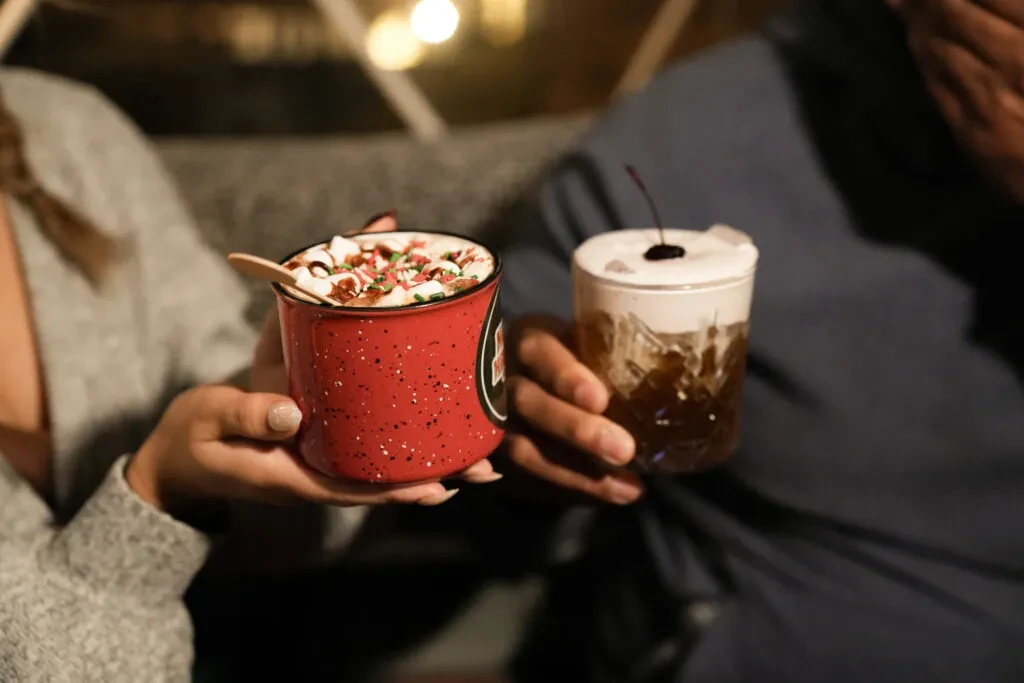 Hands holding a marshmallow hot chocolate and a cherry cocktail.