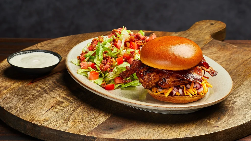 Brioche burger with melted cheese, green salad, and dip on a wooden board.