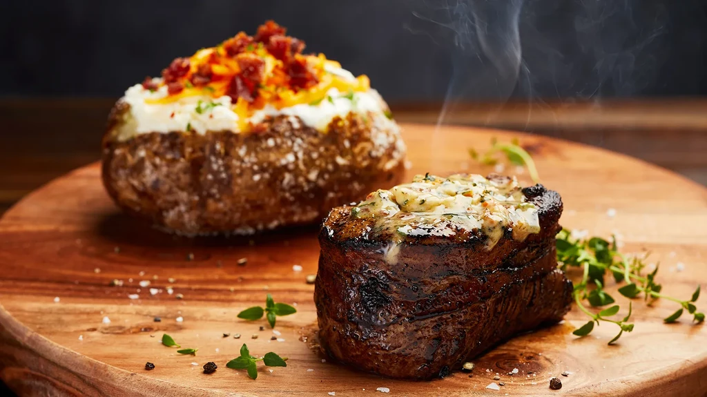 Steaming seared steak, loaded baked potato, on a wooden board.