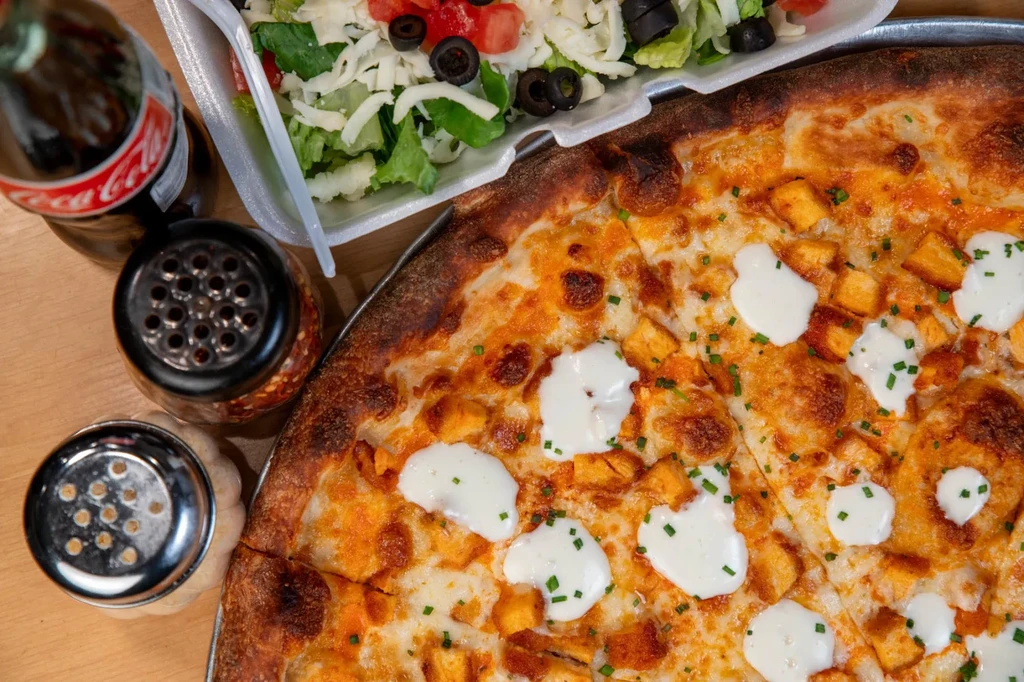 Buffalo chicken pizza, side salad, Coca-Cola, and condiments on a wooden surface.