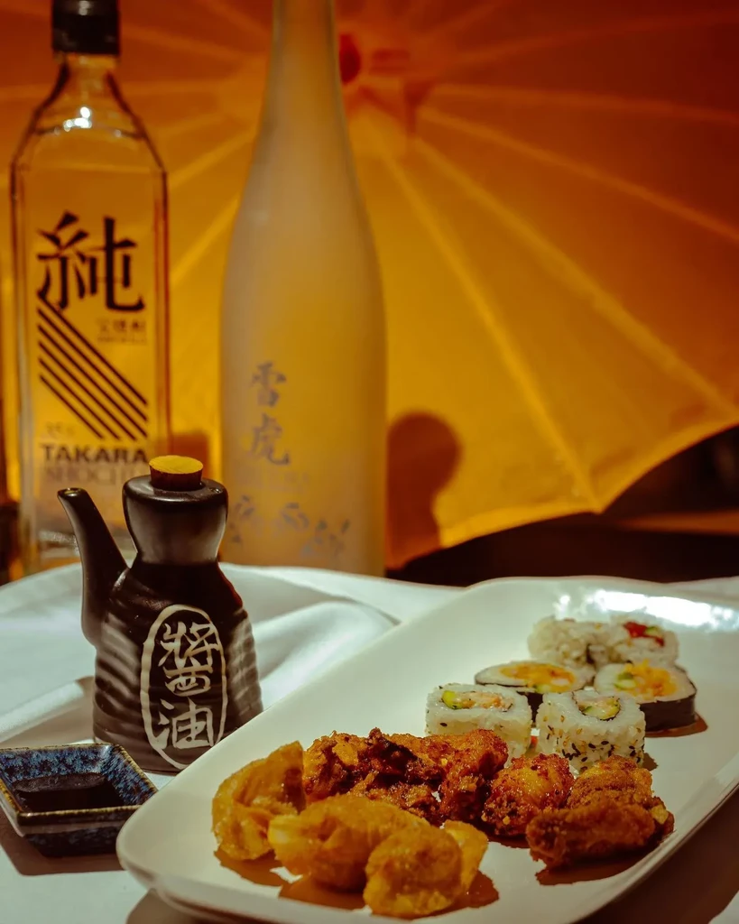 Plate of sushi, fried items, shochu, and a Japanese umbrella backdrop.