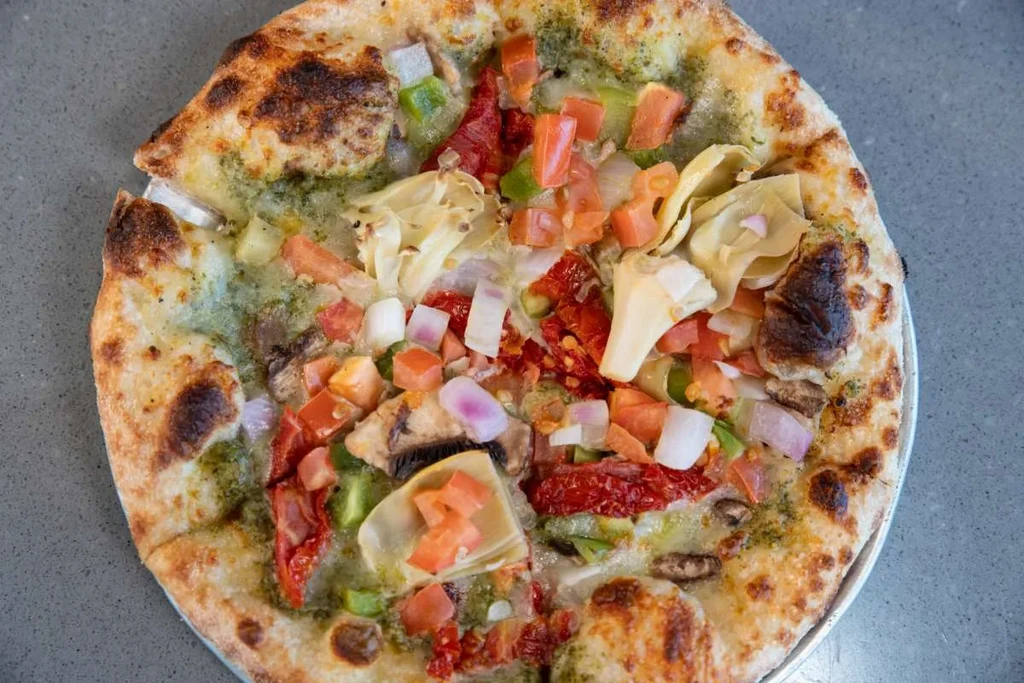 Overhead view of a fresh pesto pizza with diverse vegetable toppings.