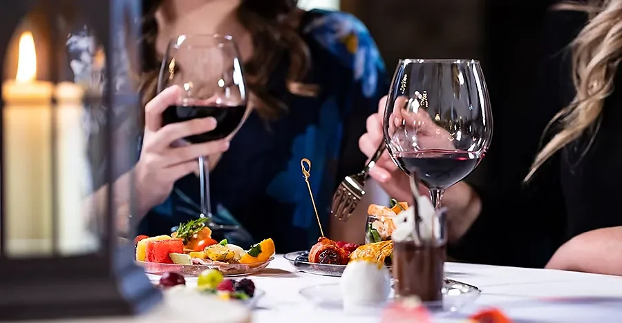 Two guests holding red wine glasses over appetizer plates at candlelit table