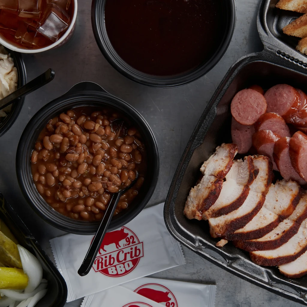Rib Crib BBQ takeout with chicken, sausage, beans, sauce, and pickles.
